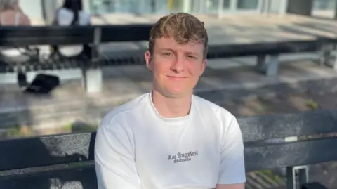 24 -year -old Oliver Reaper, Cardiff, on a bench in Center Square, outside the bus stop. They have short curly blond hair and in the gothic scenario 'Los Angeles' wearing a white T -shirt, holding her left arm with her right hand. The image focuses on him, but there are a few women on a bench behind him, the background is blurred.  