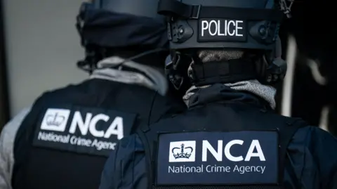 Two officers from the National Crime Agency are seen from behind with NCA visible on their black and grey uniforms. They are wearing black helmets with "POLICE" on them in white lettering
