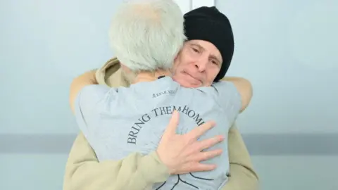 Reuters Released American-Israeli hostage, Keith Siegel, wearing a hat, embraces a loved one, the back of whose shirt reads "Bring them home" in an Israeli hospital