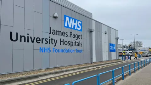 Andrew Turner/BBC The side of the James Paget Hospital building, in large lettering it says "NHS James Paget University Hospitals NHS Foundation Trust". On the right are ambulances.