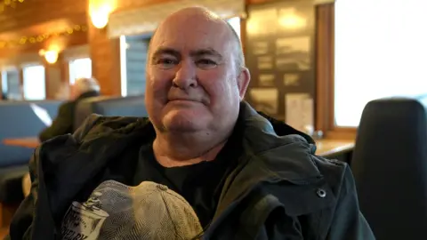 Jonathan Swingler/BBC Tony Swift has a bald head and is wearing a dark jacket over a dark t-shirt with a white design on it