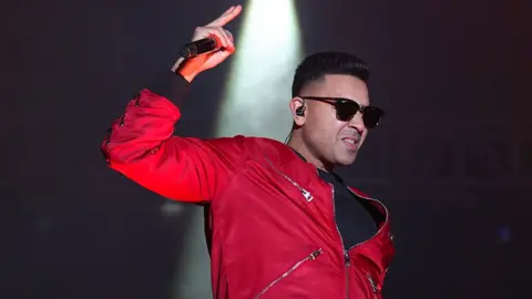Jay Sean, a man holding a microphone on stage, wearing a red fleece and sunglasses
