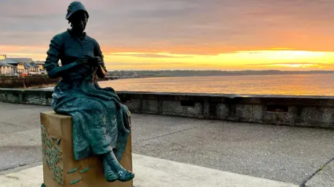 octopock A bronze statue of a woman knitting sits on a stone plinth on Bridlington's North Pier. Behind the statue the sun is low in the sky above the sea.