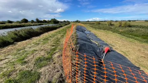 Black tarpaulin covers a long stretch of damaged river bank. The section is fenced off with temporary, orange netting. To the left, a river can be seen cutting across a grassy landscape. The sky is blue with white clouds. 