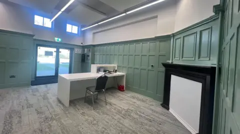 Room with green wooden panels, plastered over black and white fireplace with desk and chair in the middle