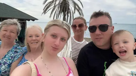 Family photo A selfie of six people in a row. In the middle is a blonde teenage girl. On the left are two older ladies in blue floral tops. On the right is a blonde woman in a striped top and sunglasses, a man with spikey brown hair, sunglasses and a black t shirt, and a young boy in a white t shirt. Behind them is a beach with the sea and a palm tree visible.