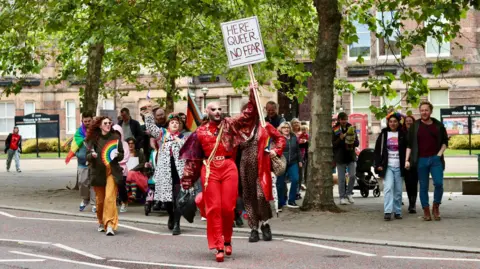 Dross Queen A group of people, some with rainbow flags, walk through St Helens led by drag queen Dross Queen dressed in red trousers and red top holding a sign that says: Here. Queer. No Fear.