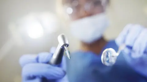 Getty Images A stock image of a dentist, who is out of focus and unidentifiable, leaning over the camera with two metal tools in their hands. The tools are made of shiny grey metal - on the right is a mirror, and on the left is a tool used for polishing. The dentist is wearing light blue scrubs and is wearing light blue sterile gloves, a face mask, and goggles. 