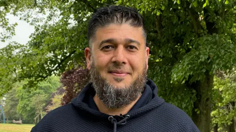 A man standing in a park. He has a black and grey beard and short black hair. He is wearing a navy hoodie and is standing in front of large trees. He is smiling into the camera.