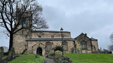 Andrew Rogers Side view of Stranton Church. The listed structure, in sandstone, has a square clock tower and there are two high arched windows in the central structure, and one in a further building to the side. A path leads up to the porch from a graveyard.