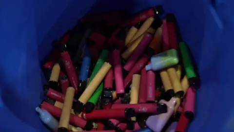 A bin full of dozens of colourful disposable vapes, mostly cylindrical but some rectangular of green, pink, yellow and lilac 