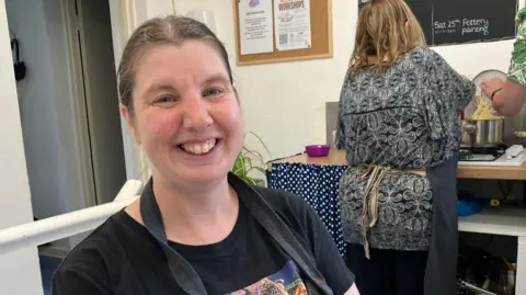 Kate Bradbrook/BBC Lady with dark hair tied back, wearing a black t-shirt and apron smiles at the camera. Behind her a ladt with brown hair and a black and white patterned top stands at the stove cooking