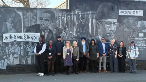 Sandwell Council Eleven people, men and women, are standing by a wall and facing the camera. The wall includes a number of large and smaller depictions of faces.
