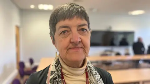 A woman wearing a white jumper and a black jacket is sat in an office. 