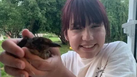 Elise Sebastian holding a small bird in her hand. She is smiling and is standing in a wooded area. She has a fringe and brown hair with a red tinge. She is wearing a white T-shirt.