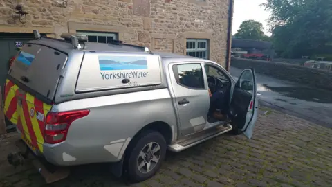 Dean Majors A silver car with Yorkshire Water branding on the side parked outside a house in Skipton with a water leak on the street outside.