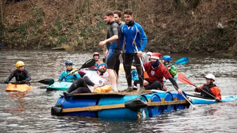Sam Hepburn A raft and five canoes are floating on a river. Three men are stood on the raft, one is laying down on it and another is sat holding an oar. People in waterproof jackets and helmets are using oars to paddle in the canoes. There is a steep leafy bank behind them.