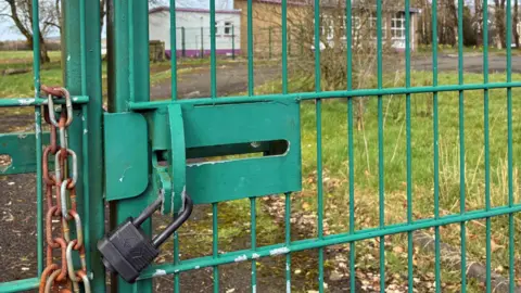 A green gate with a locked black padlock. Behind the gate is a small building with grass and trees around it. 