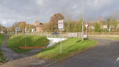 Google A road on the right curving round to the left, and entering a roundabout. There are railings either side of the carriageway on the roundabout. There is a white 40 Zone ENDS sign in the foreground and a Give Way sign beyond that. There is a sloping pavement on the left which enters a white walled underpass beneath the roundabout.