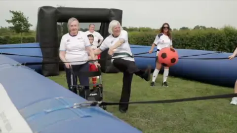 Two female care home residents taking part in a game called human table football they are inside an inflatable games course and are attached to one another with bungee cords. There are people in the background and one of the women is kicking a red football.