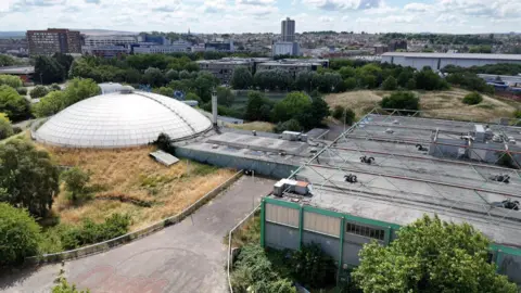 A drone shot of the Oasis site: the dome is on the left, surrounded by overgrown greenery. on the right, a large hall building. Swindon stretches out into the distance