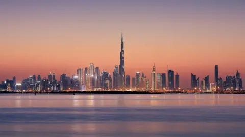 Getty Images A city skyline. In front of it is a very flat body of water, while tall skyscrapers are in the distance. It is sunset. 