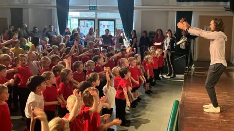 Image of a room filled with young schoolchildren, wearing red tops and black shorts and trousers. They are all looking at a stage, with their arms in the air, copying a man with curly hair who is wearing a grey hoodie, black trousers and white trainers. In the background can be seen a number of adults standing against a large window watching.