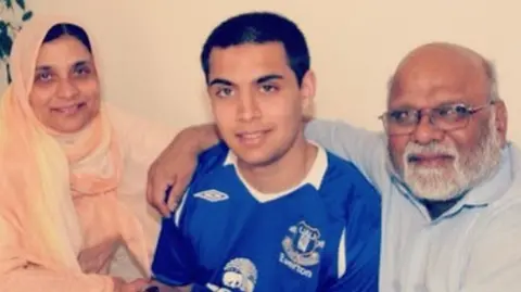 Nishat Siddiqi A smiling woman wearing a white top and peach coloured headscarf and a bearded man with glasses wearing a blue shirt hug a young black-heared boy wearing with a blue Everton football top 