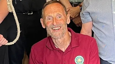 Mike Wigney looks and smiles at the camera. He is an older man with short brown hair and a beard.