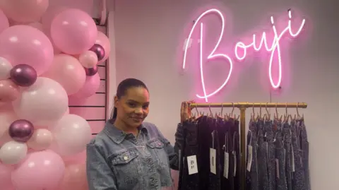 BBC/Alex James A woman is standing between a pink balloon arch and a rack of sparkly jeans. She is wearing a denim jacket top and is wearing her black hair slicked back. On the wall behind her is a pink neon sign that says Bouji. 