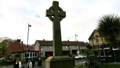 Ged Parker A large war memorial is in the centre, with railings around it. Behind is a row of housing.  