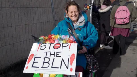 Melissa Topping, holding a sign which reads "stop the EBLN" - which stands for East Bristol Liveable Neighbourhood. She is wearing a blue coat. 