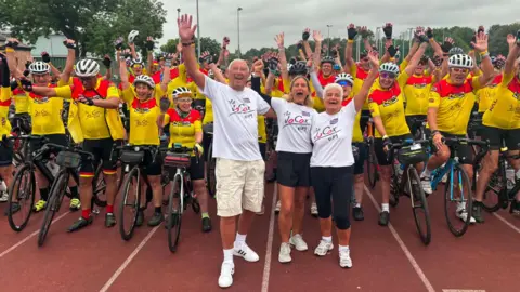 Cyclists lined-up at the start of their bike ride. They are in an athletics stadium, where the ground is red, with lanes painted in white. The cyclists are wearing yellow and read T-shirts. Standing in front are Jo Cox's parents and sister, Kim Leadbeater, who are wearing white "Jo Cox Way" T-shirts.