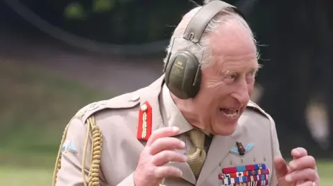 King Charles III. He is dressed in the number four tropical dress uniform which is beige. He is wearing a set of khaki green ear defenders. He is looking to the side and smiling.  