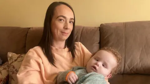 Shauna has shoulder length brown hair and is looking at the camera smiling. She is wearing a light orange sweatshirt. She is holding Harry in her arms, he has curly ginger hair and is wearing a grey jumper.