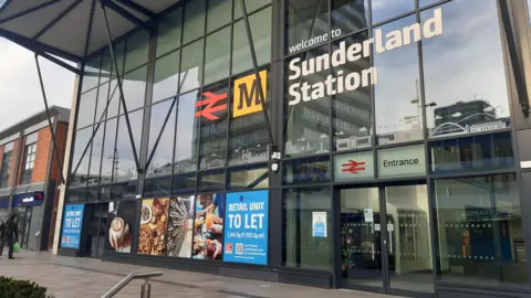 Sunderland Station is a tall glass building with sliding doors. A large sign above the entrance reads Welcome to Sunderland Station. The Tyne and Wear Metro's yellow logo is next to it. Advertising posters cover the bottom windows showing photos of coffee and food and reading Retail Unit To Let. 