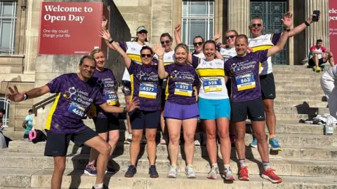 11 people stood on some city hall stone steps, waving at the camera and smiling. They are wearing purple Leeds Hospitals Charity t-shirts and running shorts and trainers.