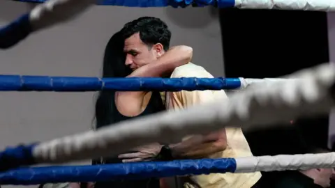 Curious North Productions Two people embrace inside a boxing ring, framed by thick blue and white ropes in the foreground. One person wears a sleeveless black top, and the other wears a light-colored shirt. The setting appears to be an indoor venue with spectators partially visible in the background.
