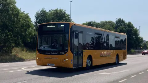 Jenny Kirk/BBC A yellow and black Sanders Coaches bus being driven along the A140 road in Norwich near to the airport on a sunny day.