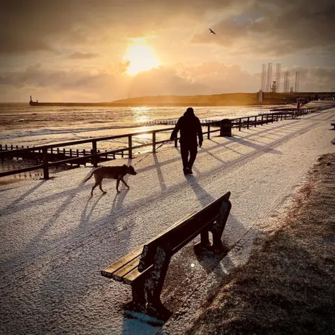 Alex Mitchell Sunrise over Aberdeen Beach. The sky is a pale yellow colour and the sun is reflecting on the water. A bench and a man walking a dog are in the foreground. There is frost on the ground.
