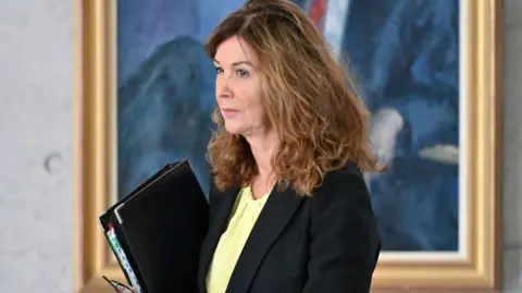 Dorothy Bain wearing a dark suit over a light green top and carrying a black folio.
