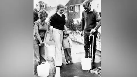 PA Wire Two women and a toddler are waiting to fill up water from a standpipe. A man had a bucket under the pipe and he has turned the tap on.