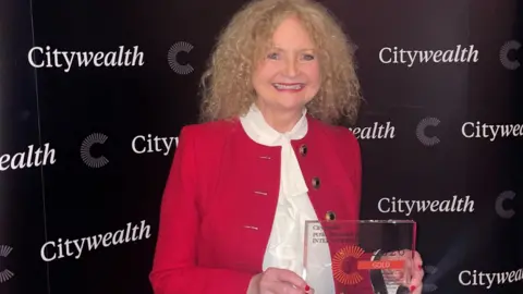 TISE A woman called Kay McCarthy holds an award she has won at a ceremony in London. She has a broad smile on her face and is wearing a red blazer with a white blouse underneath. McCarthy has long tight blonde curly hair. A sign behind her says "Citywealth". The award is mainly made from glass.
