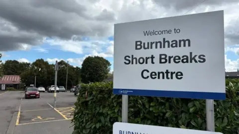 Martin Heath/BBC A large sign saying "Welcome to Burnham Short Breaks Centre" in black lettering on a white background. There is a hedge behind the sign, and a car park with yellow lines painted on the ground behind. A single-storey building with a pitched roof is visible on the left in the background.