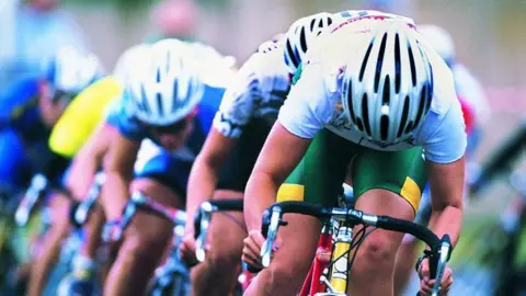 Three male cyclists with white tops and green shorts on a bike racing behind each other. All are wearing white cycling helmets.
