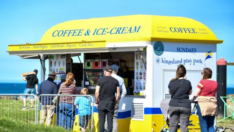 A line of people with their backs to the camera are queuing outside an ice cream stall. The stall has a yellow roof and says coffee and ice cream in blue letters. The rest of the stall is white, yellow and blue. The sun is shining, there is a blue clear sky and the sea can be seen in the distance.