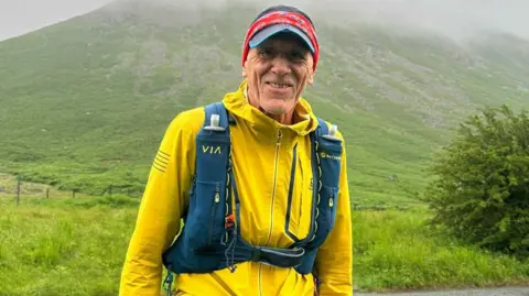Peter Rielly Ultramarathon runner Kevin Munt stands in front of a green luscious hill. He is wearing trail running clothes with a bright yellow fleece and a navy running backpack.