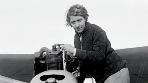 Black and white photograph of Amy Johnson dressed in flying gear in the cockpit of an aircraft. She is looking towards the camera and smiling, with her goggles pulled up on her forehead.