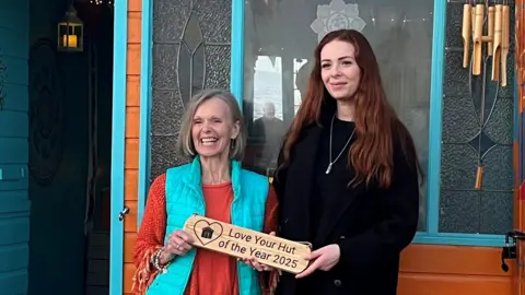 Chrissie Bond is wearing a turquoise bodywarmer above an orange top. She is smiling and holding a wooden plaque that says "Love Your Hut of the Year 2025". The plaque is also being held by Alexandra Sandberg, who is wearing all black. Behind them is the orange beach hut, which has turquoise features.