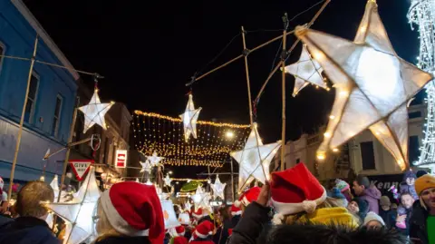 Mid & East Antrim Borough Council A large group of people wearing red and white Santa style hats. They are standing under giant star lights.